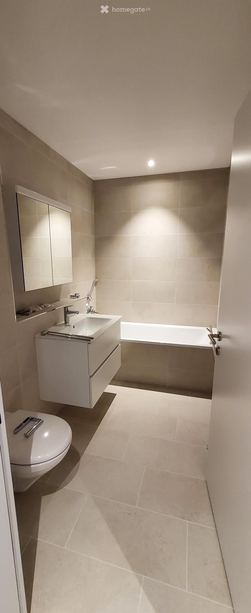Modern bathroom with beige tiles, a white sink, a bathtub, a toilet, and a mirrored cabinet.
