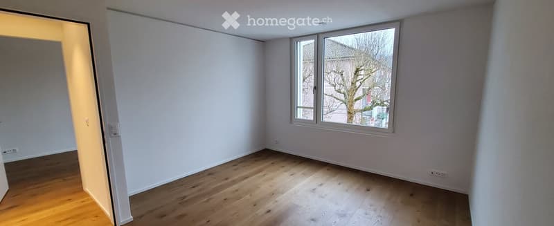 Empty room with light wooden flooring, white walls, and large windows offering a view of a tree outside. The room is unfurnished and has an open doorway leading to another space.