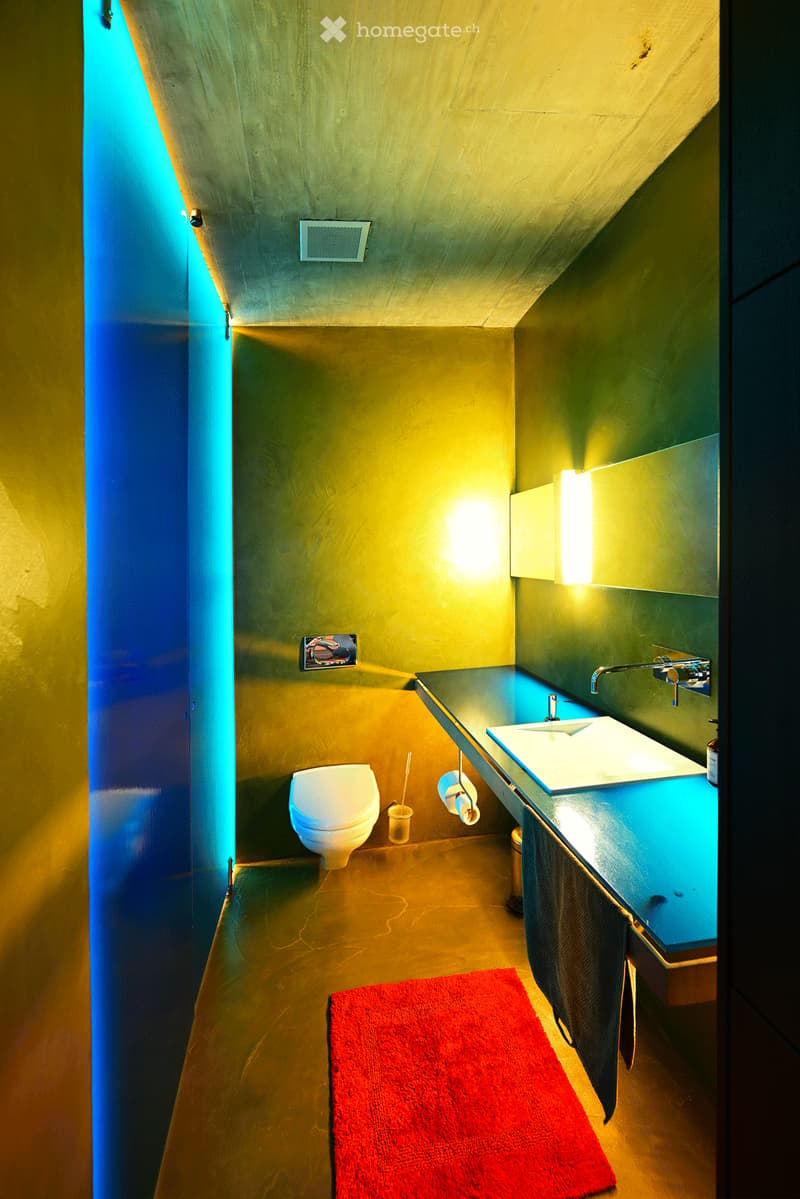 Modern bathroom with a toilet, sink, mirror, and a red rug on the floor. The walls are painted in a gradient of yellow and blue, and the lighting is warm and soft.