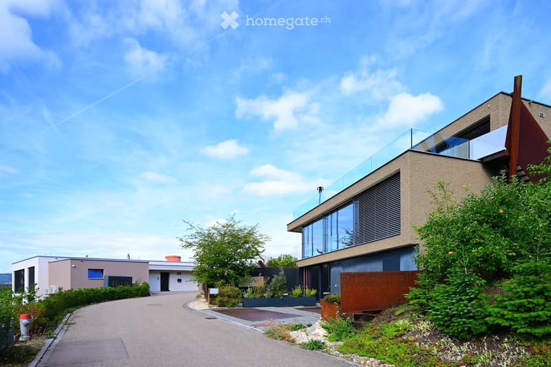 Modern two-story house with a glass balcony, surrounded by greenery and a paved driveway.