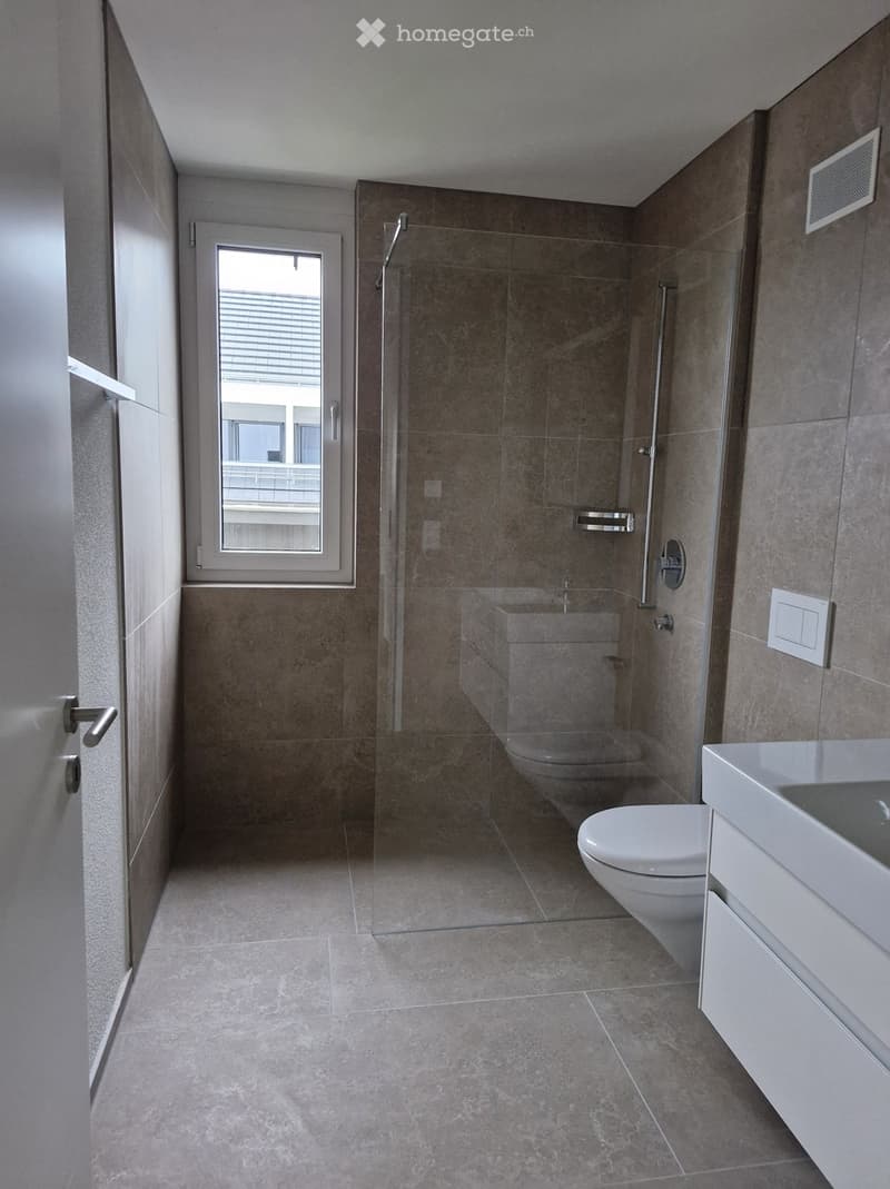 Modern bathroom with a glass-enclosed shower, toilet, and sink. The space features tiled walls and floor, a window with blinds, and a sleek, contemporary design.