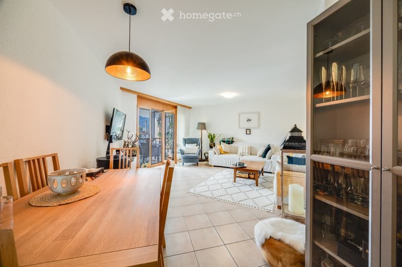 Bright living and dining area with a white sofa, coffee table, dining table with chairs, and a glass cabinet displaying glassware. The space features a modern pendant light, a floor lamp, and a view through large glass doors to a balcony.