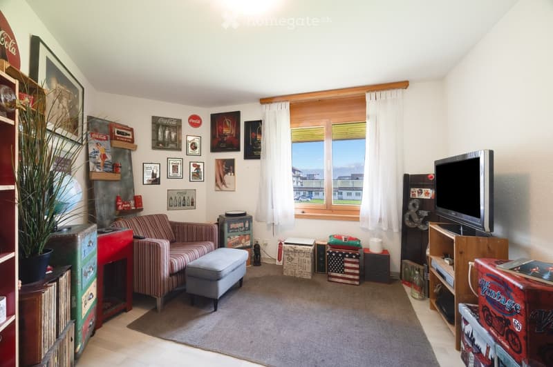 A cozy living room with a striped sofa, ottoman, television, and various decorative items including framed pictures and a plant. The room features a fireplace and large windows with white curtains.