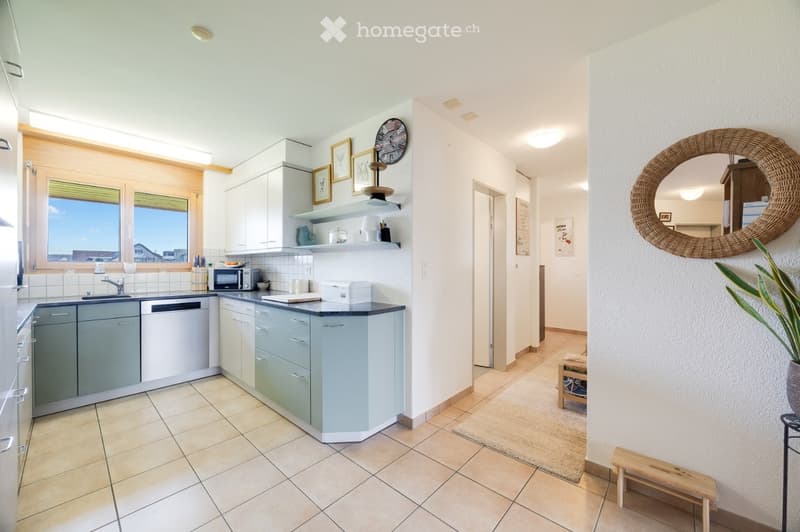Modern kitchen with light grey cabinets, white countertops, and a tiled floor. Features a sink, microwave, dishwasher, and a round woven mirror on the wall. Shelves hold decorative items and a clock. A small wooden bench and potted plant are near the entr