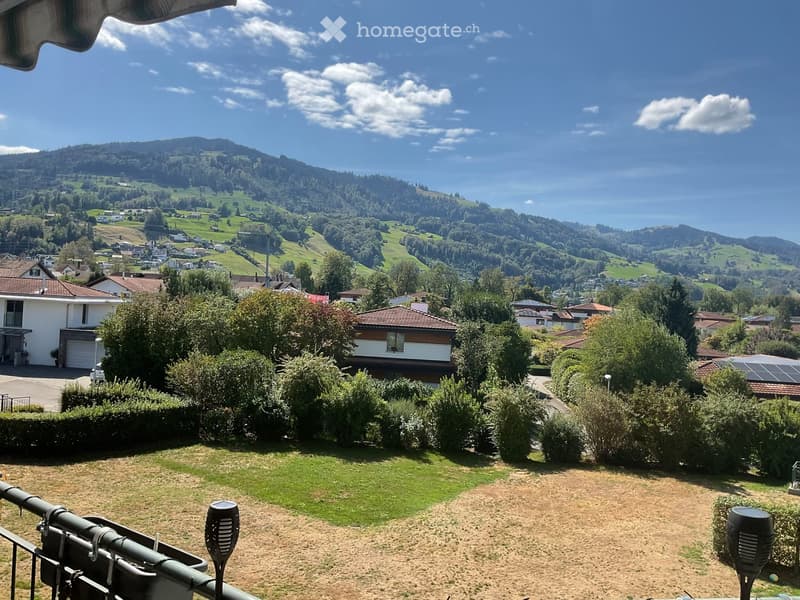 A scenic garden area with a panoramic view of a lush, green valley and distant mountains under a clear blue sky.