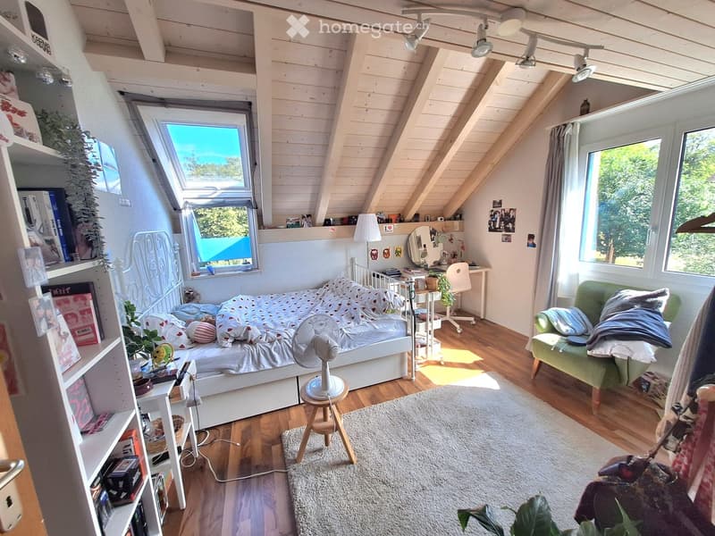 Cozy attic bedroom with wooden floors, slanted ceilings, large windows, a bed with a white metal frame, a green sofa, a small table with a fan, shelves with books and decor, and a desk area with a chair and mirror.