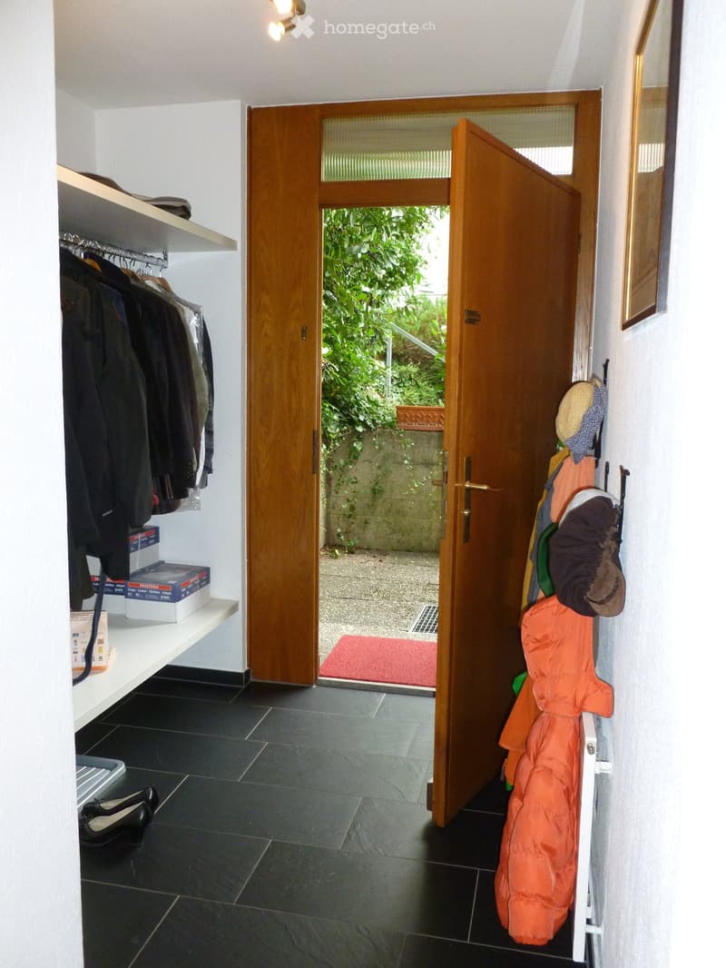 Hallway with black tiled floor, open wooden door leading to outdoor area, coat rack with jackets and hats, shelf with boxes and shoes.