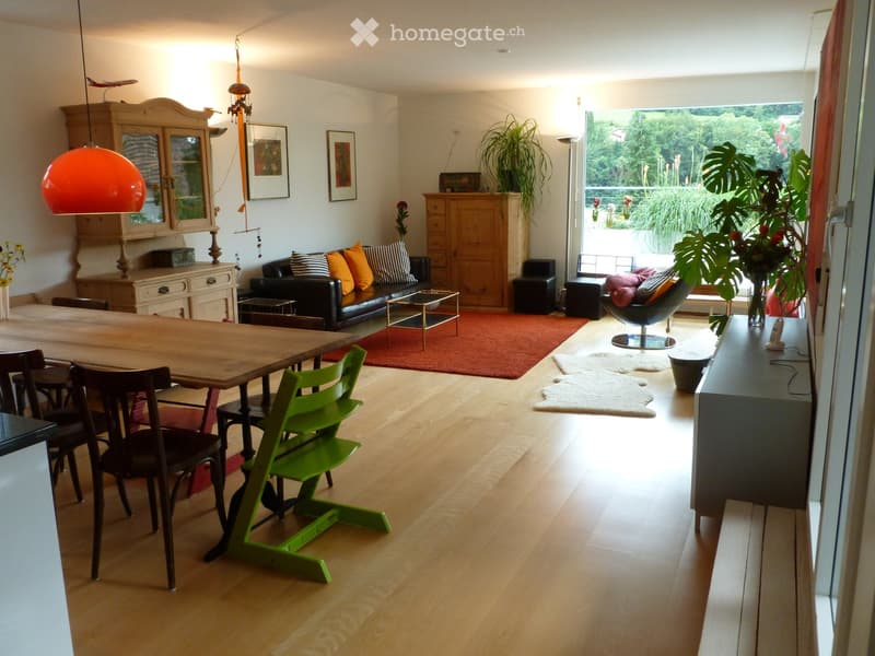 Bright living and dining area with wooden flooring, a large window, a black leather sofa with colorful cushions, a wooden dining table with chairs, a green chair, a cabinet, and decorative plants.