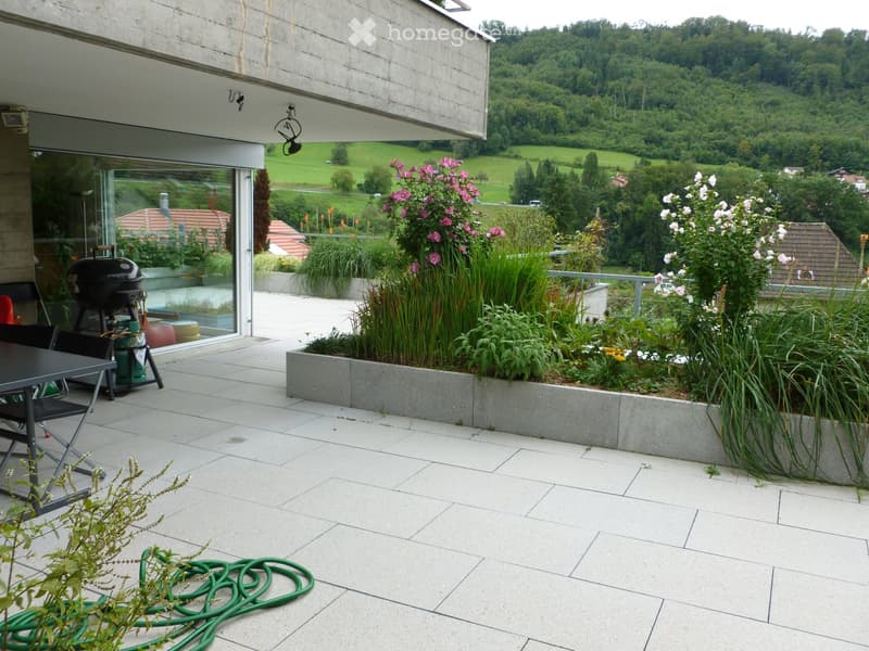 Balcony with tiled floor, modern furniture, grill, and lush plants, overlooking a scenic valley.