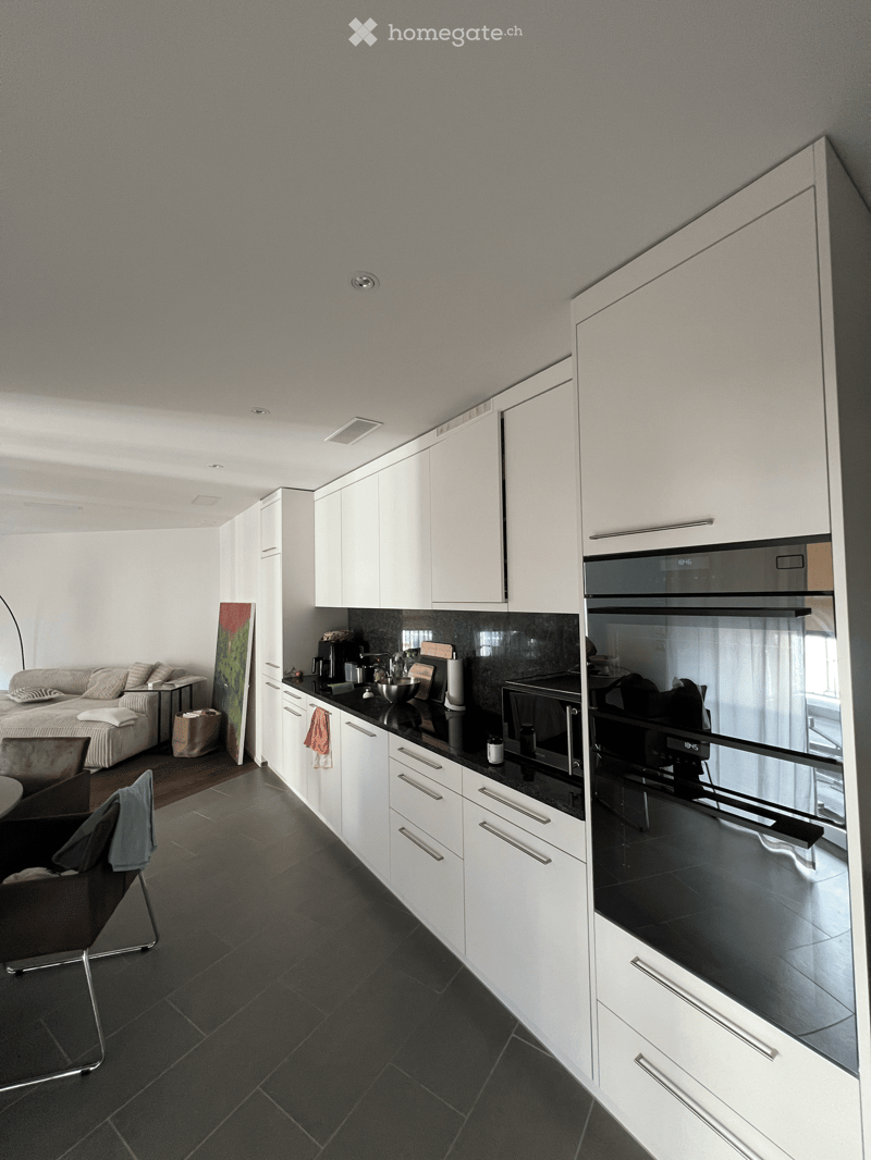 Modern kitchen with white cabinets, black countertops, and stainless steel appliances. A sofa and chair are visible in the adjacent living area.