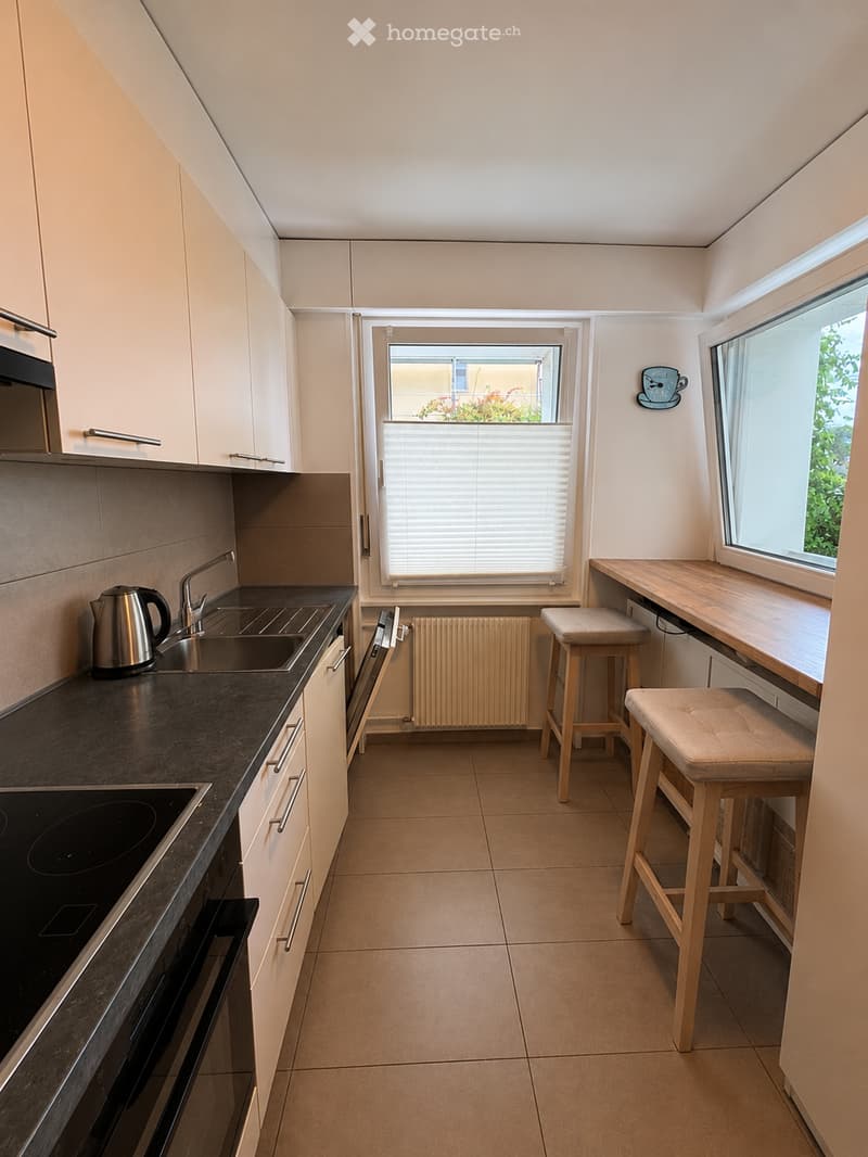 Modern kitchen with white cabinets, dark countertops, stainless steel appliances, and a small dining area with two stools and a table.