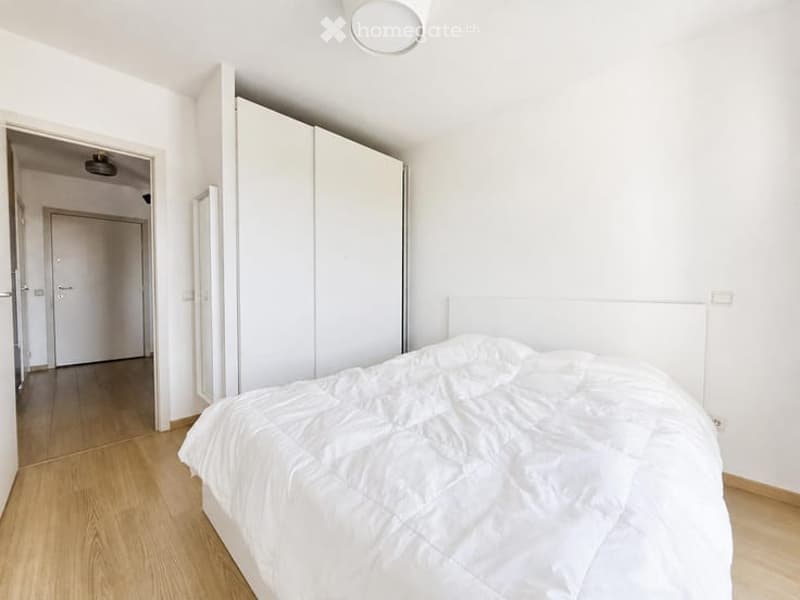 Bright bedroom with white walls, wooden flooring, a neatly made bed with white bedding, and a large white wardrobe.