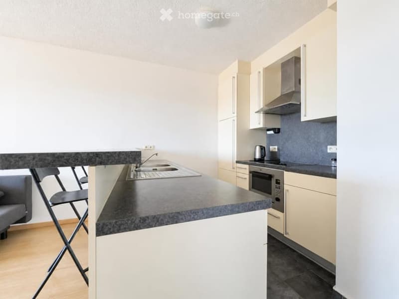 Modern kitchen with light-colored cabinets, a black countertop, and stainless steel appliances including a sink, stove, and oven. A coffee maker is placed on the counter, and a chair is visible near the island.