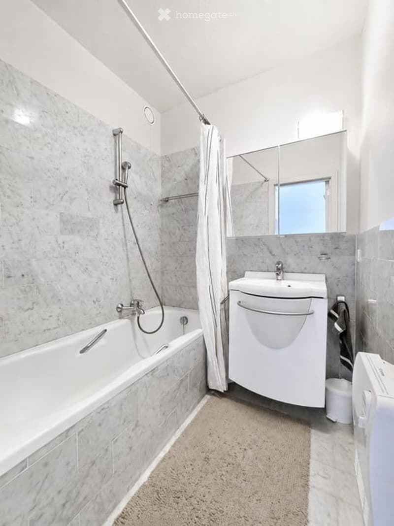 Modern bathroom with a white bathtub, shower curtain, sink, mirror, and cabinet. The walls are tiled in a light grey marble pattern, and a small towel is visible on the floor.
