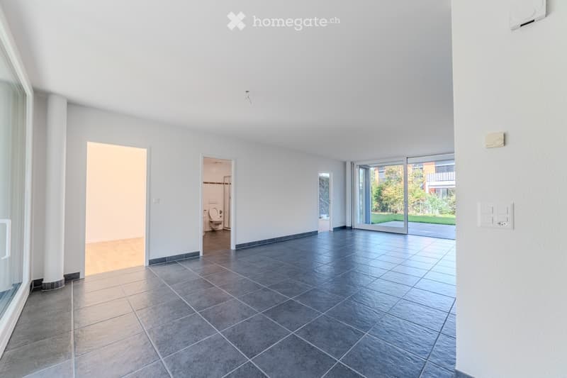 Empty room with grey tiled floor, white walls, large windows, and a view of the garden. No furniture or equipment present.