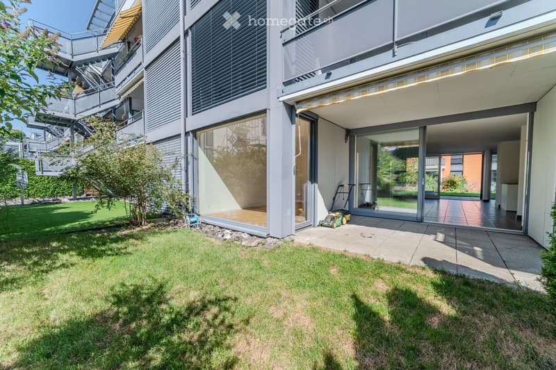 Modern residential building with a well-maintained garden area, featuring a lawn, small trees, and a paved patio with large glass doors leading to the interior.