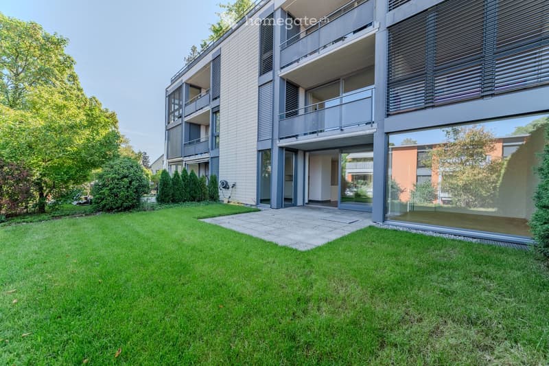 Modern multi-story building with balconies, large windows, and a well-maintained lawn in front.