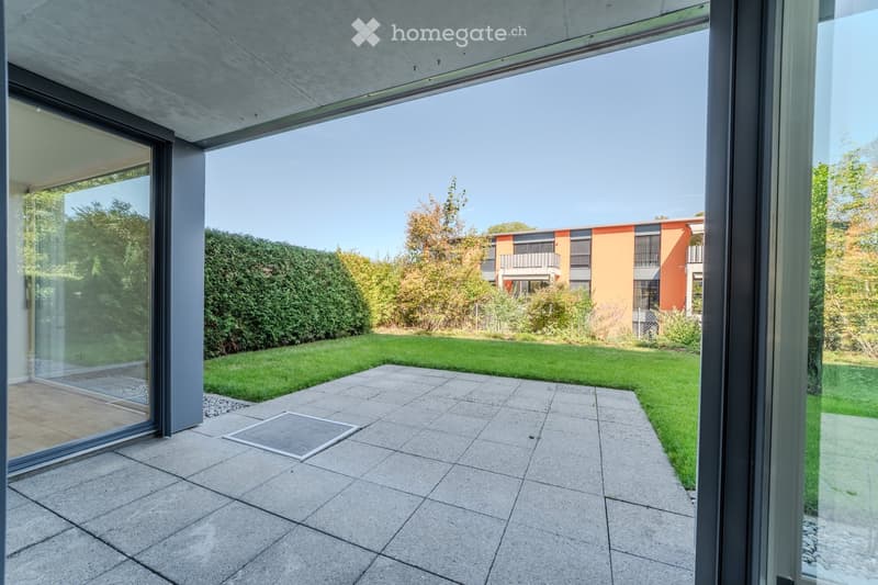 Modern outdoor patio with large glass doors leading to a well-maintained lawn and surrounding greenery, with a contemporary building in the background.