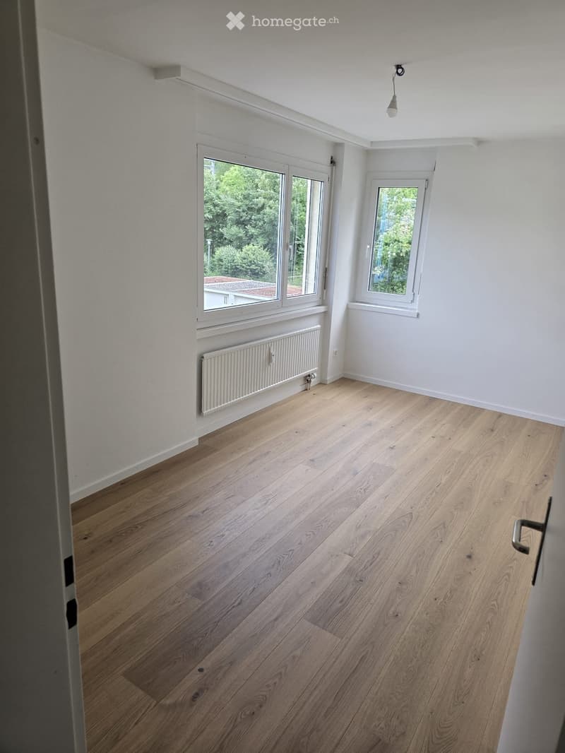 Empty room with light wooden flooring, white walls, and large windows offering a view of greenery outside. A radiator is visible under one window.