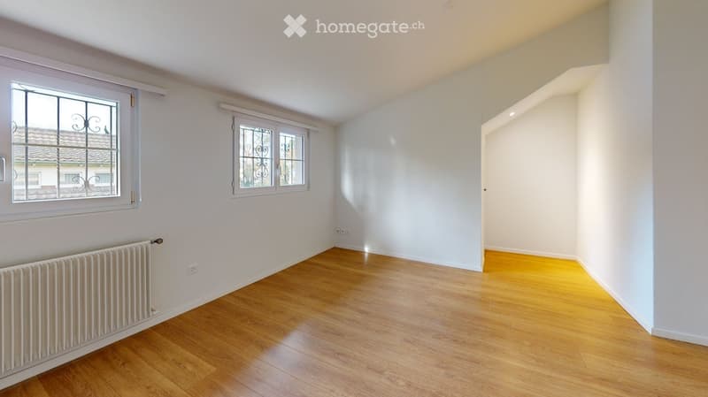 Empty room with wooden flooring, white walls, and two windows with decorative metal grilles. Radiator visible on one wall.