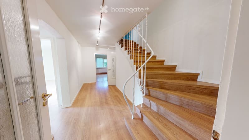 Spacious hallway with wooden flooring, white walls, and a staircase with wooden steps and a white metal railing. A door is visible on the left, and a room with a window is visible at the end of the hallway.