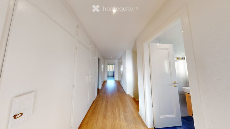 A long, narrow hallway with white walls, wooden flooring, and several white doors on both sides. A small bathroom is visible at the end of the hallway.