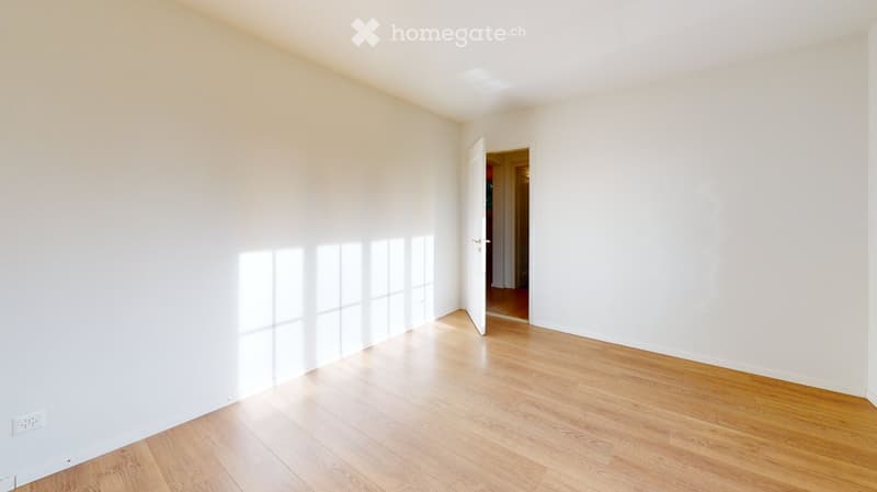 Empty room with white walls and wooden flooring, featuring a large window and an open doorway leading to another room.