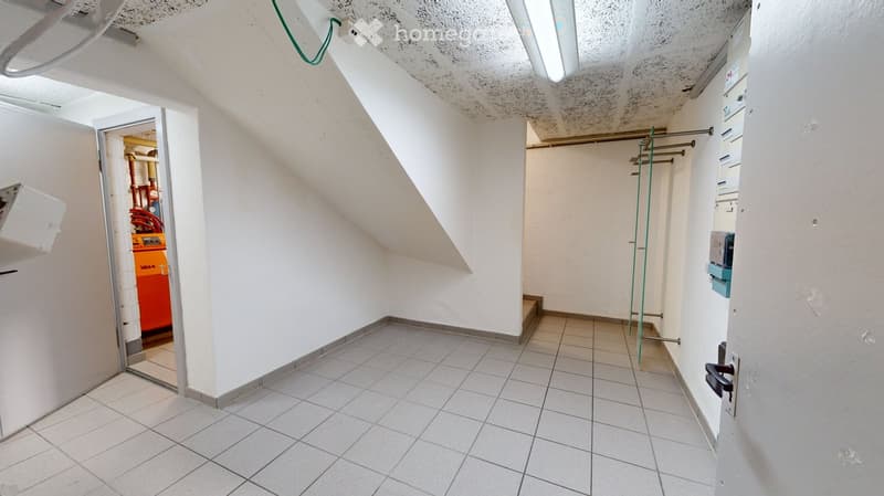 Empty hallway with white walls, tiled floor, and exposed pipes in the background. The ceiling has visible wiring and a light fixture.