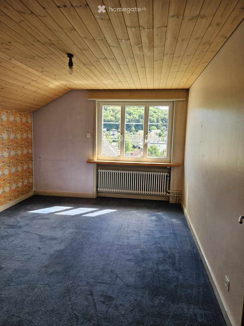 Empty room with blue carpet, wooden slanted ceiling, large windows showing outdoor view, radiator, and patterned wallpaper on one wall.