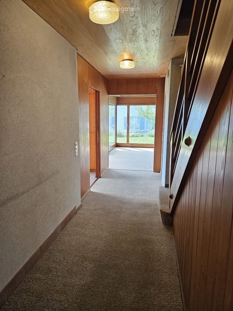 Carpeted hallway with wooden panel walls, recessed lighting, and a glass door leading outside.