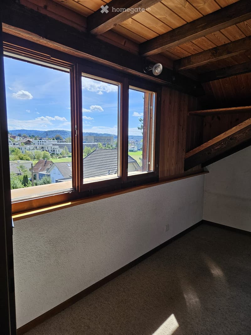 Empty room with wooden ceiling beams, large windows offering a view of the town and surrounding landscape, and a wooden staircase leading upwards.