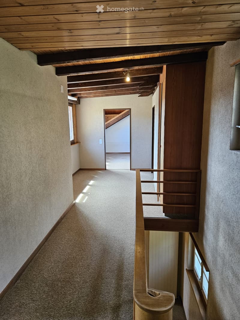 Spacious hallway with wooden ceiling beams, carpeted floor, and wooden staircase leading upwards. Natural light from side windows illuminates the area.