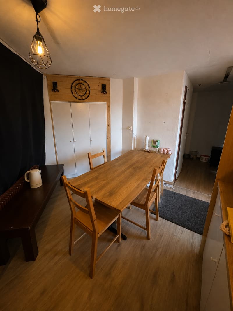 Bright kitchen with wooden flooring, a wooden dining table surrounded by chairs, white built-in cabinets, and a teapot on a sideboard.