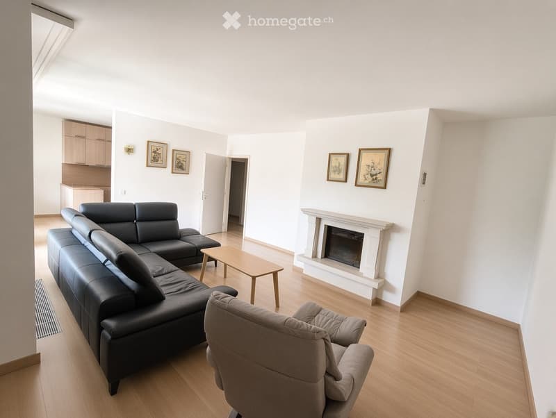 Modern living room with light wooden flooring, white walls, a black sectional sofa, a wooden coffee table, a grey armchair, and a white fireplace with framed pictures above it.