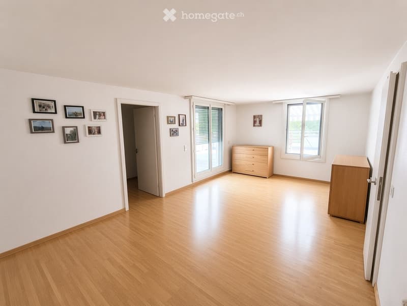 Empty room with light wooden flooring, white walls, and large windows with blinds. There are several framed pictures on the walls and a sliding glass door leading to a balcony area.
