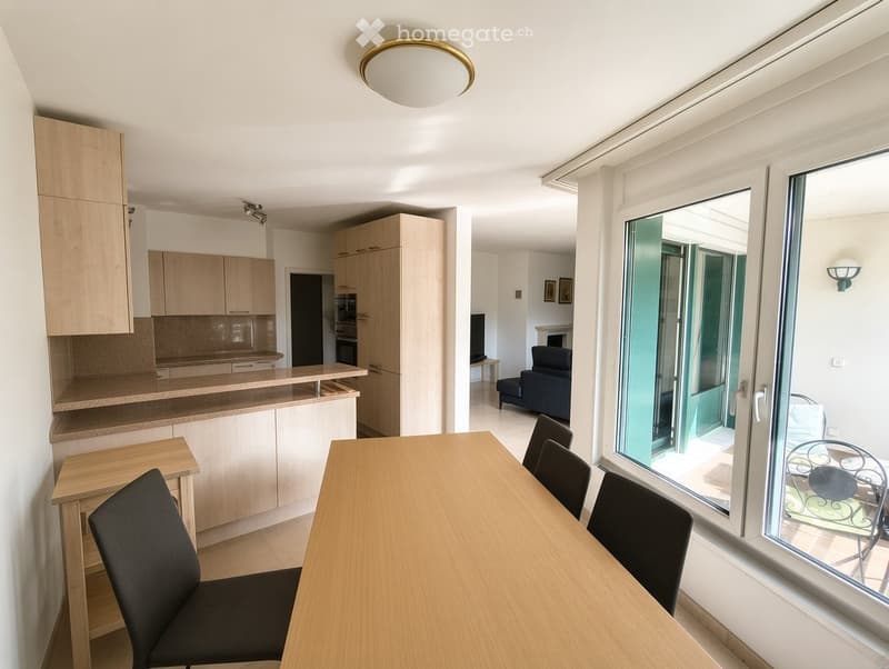 Modern, well-lit room with a dining table, chairs, a sofa, and a kitchen area with cabinets and an oven. Large windows and a balcony door provide natural light and access to the outdoor area.