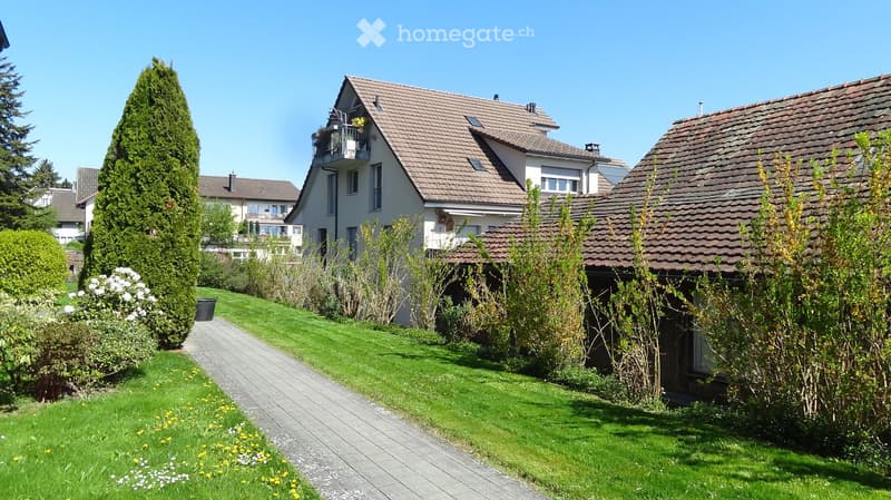 Garden / Outdoor Area. A well-maintained garden area with a paved pathway leading to a house, surrounded by green grass, shrubs, and trees. The house features a tiled roof and multiple windows, with a small balcony visible on the upper floor.