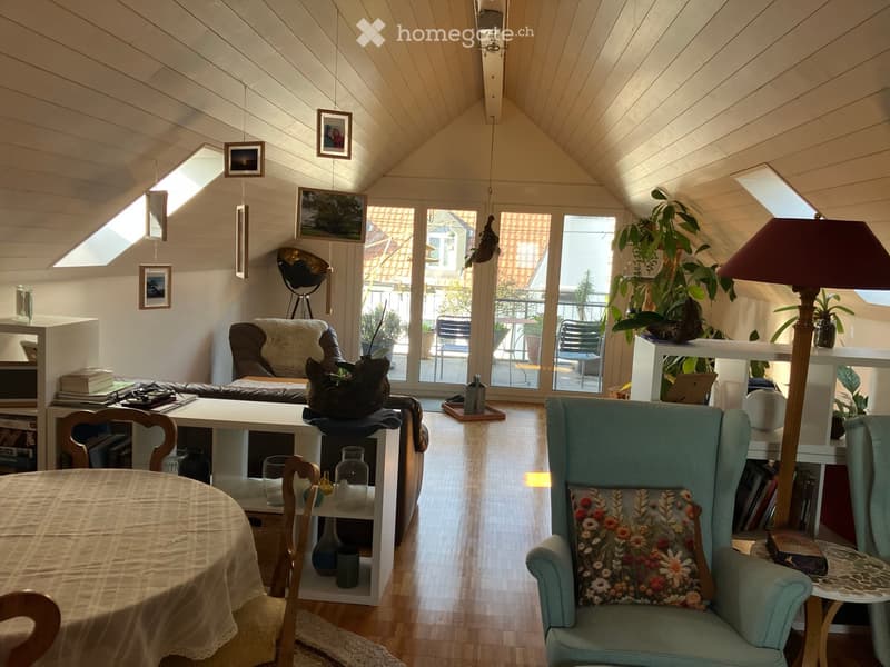 Furnished Room. Bright, airy room with wooden floors, large windows, and skylights. Features a green armchair, sofa, dining table, shelves, and decorative items.