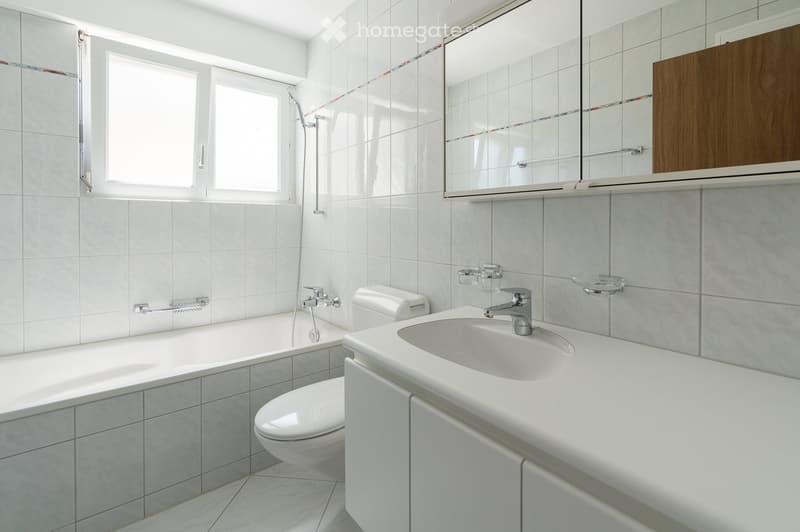 Modern bathroom with white tiles, bathtub, toilet, sink, mirror, and showerhead.