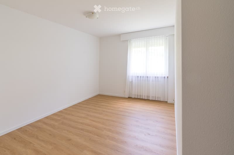 Empty room with light wooden flooring, white walls, and a window with white curtains.