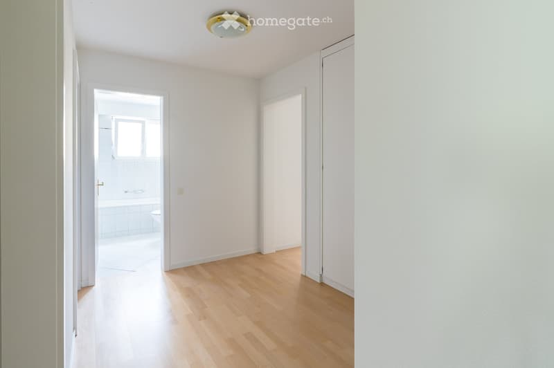 Empty hallway with light wooden flooring, white walls, and a ceiling light fixture. Two doors lead to other rooms, one of which is a bathroom.