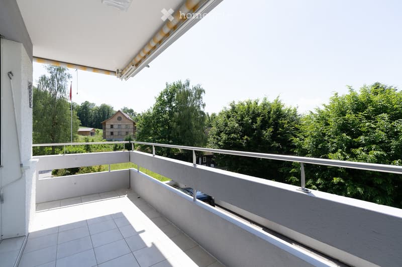 Balcony with tiled floor, metal railing, and retractable awning, overlooking a green landscape with trees and a small house.