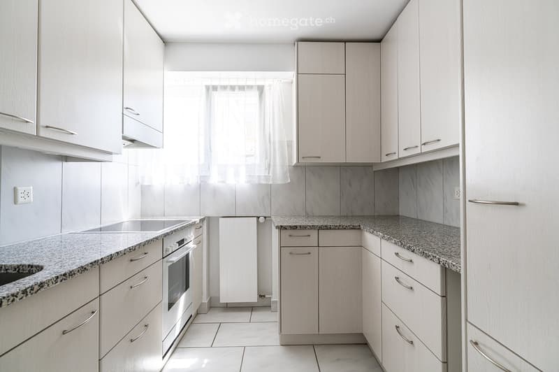 Modern kitchen with light wood cabinets, granite countertops, and stainless steel appliances including a sink and oven. The space is bright with natural light from a window.