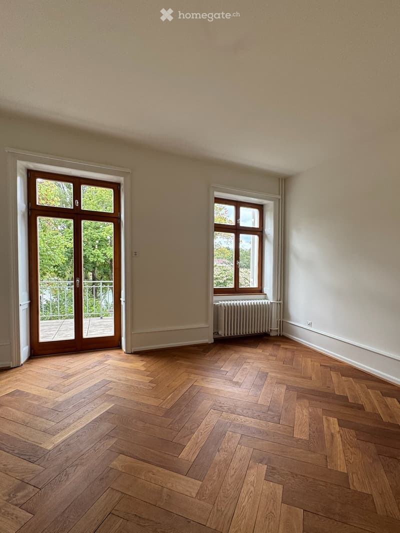 Empty room with wooden herringbone flooring, large window with radiator, and sliding glass door leading to a balcony.