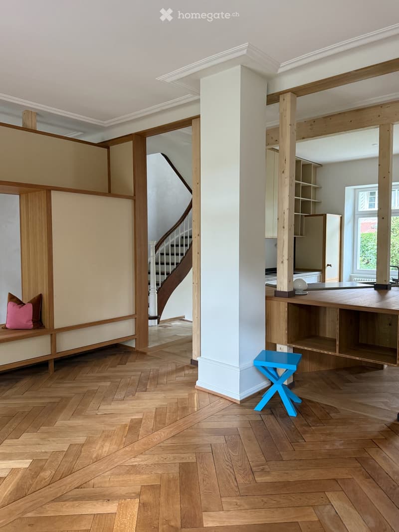 Open-plan living area with herringbone wooden flooring, built-in bench seating, a small blue stool, a central table, and visible staircase leading upwards.