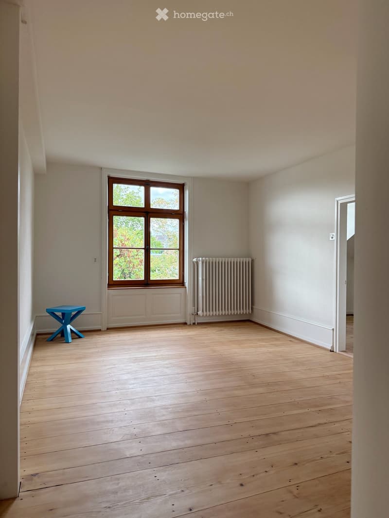 Empty room with wooden flooring, light-colored walls, a window with wooden frame, radiator, and a small blue stool.
