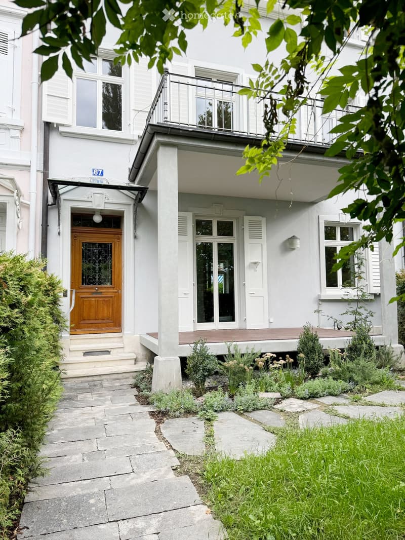 Two-story white building with a wooden front door, balcony, and small garden area with plants and shrubs.