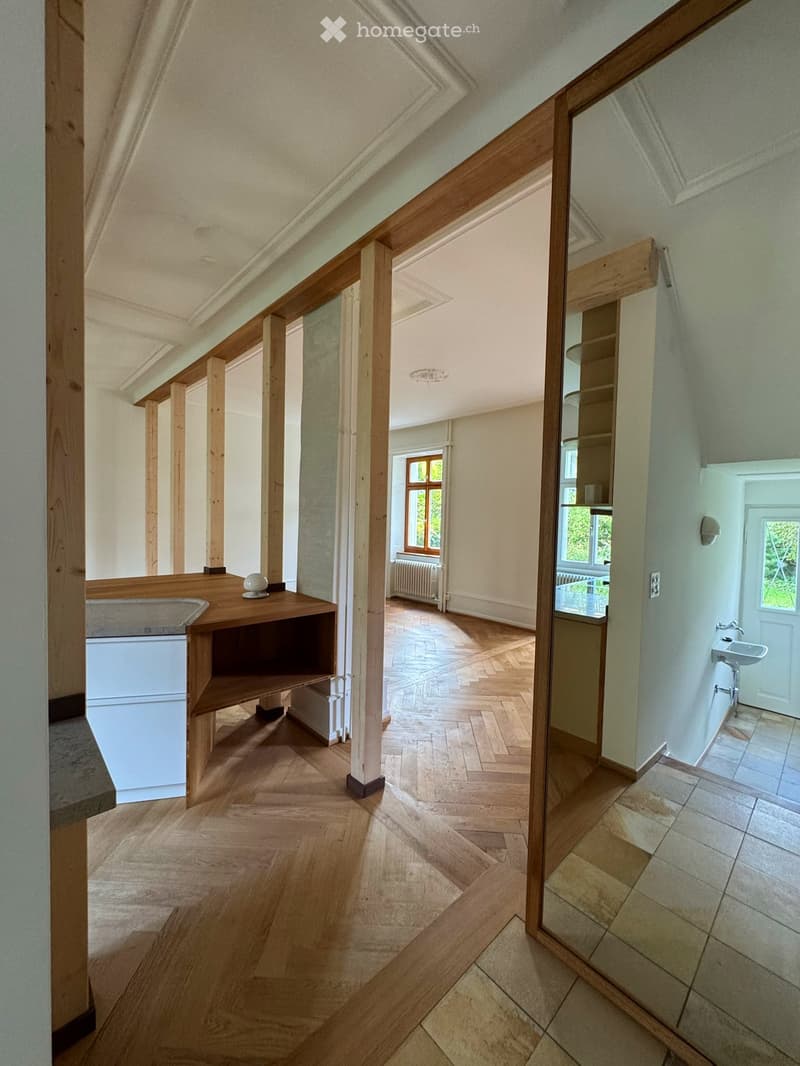 Spacious hallway with wooden flooring, wooden beams, and a small wooden shelf with a decorative item. The hallway leads to a room with a sink and a window.
