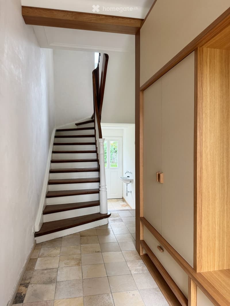 Spacious hallway with wooden staircase, white walls, tiled floor, and a small sink visible in the background.