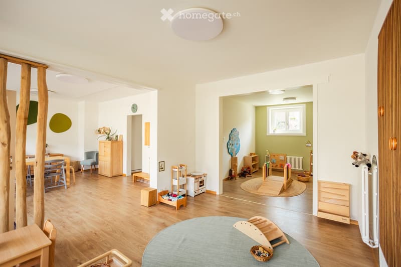Bright, open-plan living and play area with wooden flooring, modern furniture, and various toys and play structures.